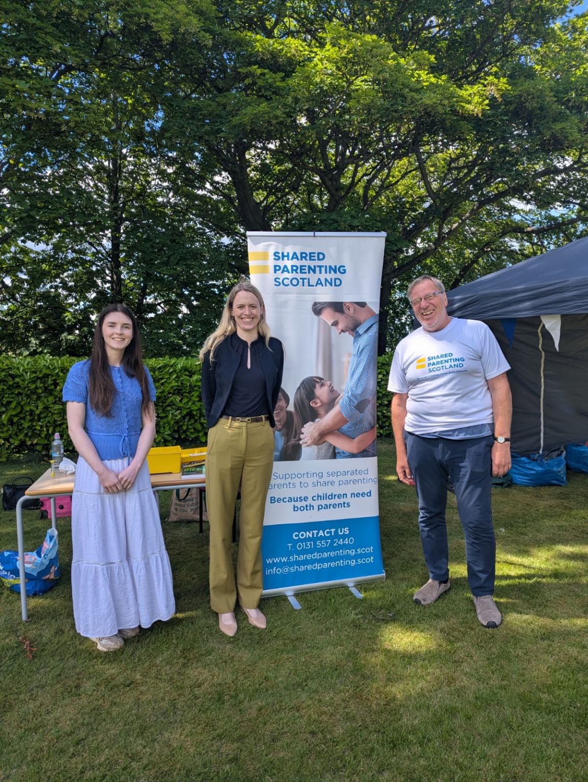 Armed Forces Big Day Out 20 June A picture of, from left to right: Megan Hutcheon, Shared Parenting Scotland; Louise Sandher-Jones MP, Armed Forces Minister; and Guy Holloway, Shared Parenting Scotland trustee, at the Armed Forces Big Day Out hosted at Edinburgh Napier University on 20 June 2026. Between Louise and Guy is a tall Shared Parenting Scotland banner with a photo of a father hugging his daughter and a message underneath saying 'Supporting separated parents to share parenting. Because children need both parents' Shared Parenting Scotland contact details are at the bottom of a banner on SPS blue background: CONTACT US T: 0131 557 2440 www.sharedparenting.scot info@sharedparenting.scot