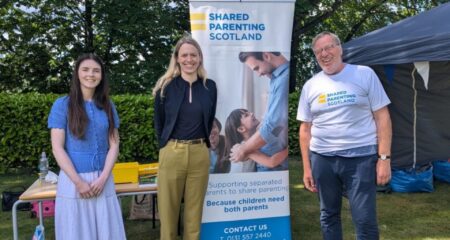 A picture of, from left to right: Megan Hutcheon, Shared Parenting Scotland; Louise Sandher-Jones MP, Armed Forces Minister; and Guy Holloway, Shared Parenting Scotland trustee, at the Armed Forces Big Day Out hosted at Edinburgh Napier University on 20 June 2026. Between Louise and Guy is a tall SPS banner with a photo of a father hugging his daughter and a message underneath saying 'Supporting separated parents to share parenting. Because children need both parents'