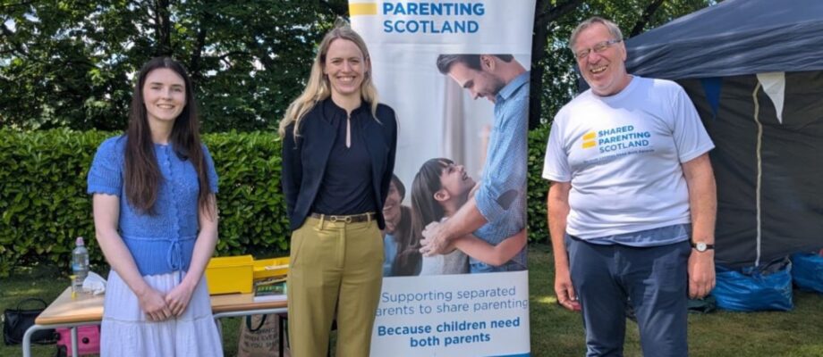 A picture of, from left to right: Megan Hutcheon, Shared Parenting Scotland; Louise Sandher-Jones MP, Armed Forces Minister; and Guy Holloway, Shared Parenting Scotland trustee, at the Armed Forces Big Day Out hosted at Edinburgh Napier University on 20 June 2026. Between Louise and Guy is a tall SPS banner with a photo of a father hugging his daughter and a message underneath saying 'Supporting separated parents to share parenting. Because children need both parents'