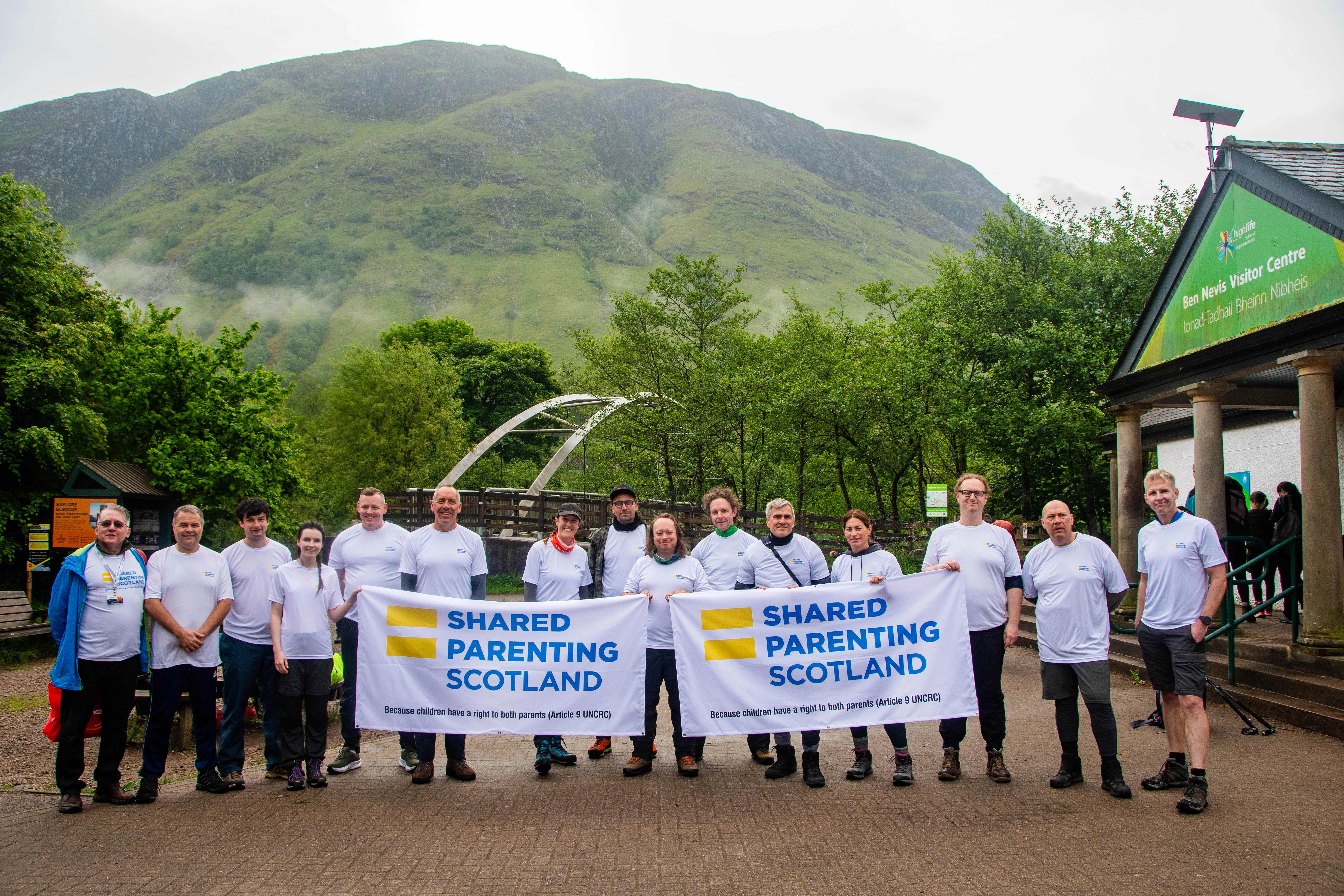 FRZ_7472 A group of 15 men and women wearing white T-shirts with the Shared Parenting Scotland logo stand outside the Ben Nevis Visitor Centre with two Shared Parenting Scotland flags. Ben Nevis is visible in the background on an overcast morning.